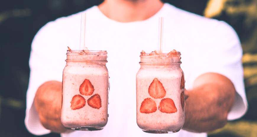 Egy srác mindkét kezében egy-egy rózsaszín smoothie-t nyújt feléd, amiben eper van.