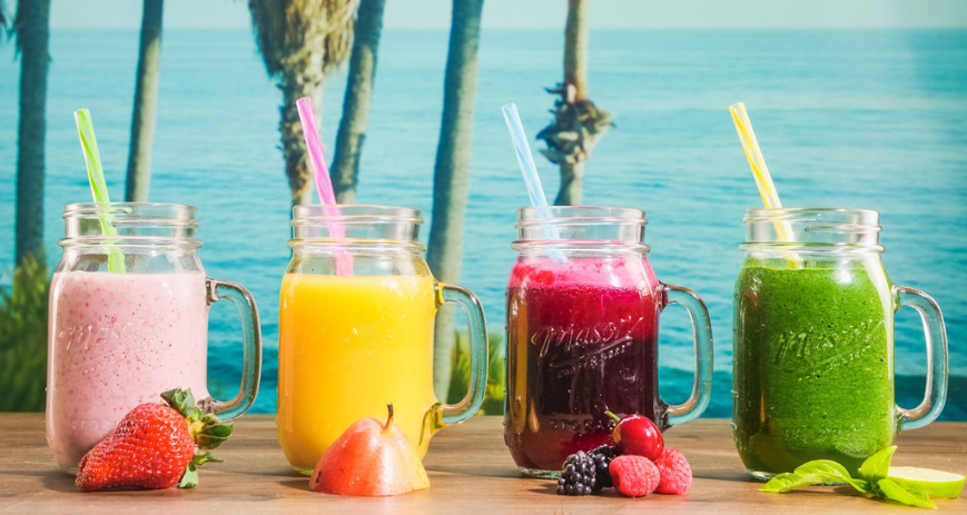 Vízpart a háttér, a képen 4 különböző színű smoothie, előtte gyümölcsökkel